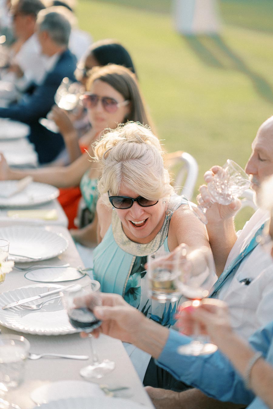 A group of people enjoying an outdoor meal, smiling and toasting with wine glasses at a long dining table during a sunny event.
