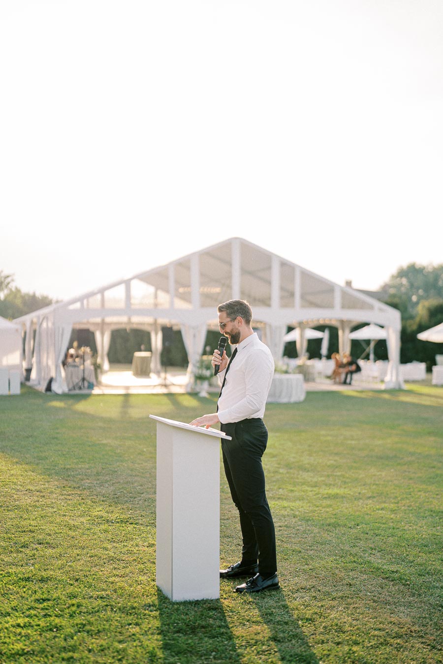 A man in formal attire giving a speech outdoors with a microphone and podium, standing in front of a large white event tent during a sunny day.