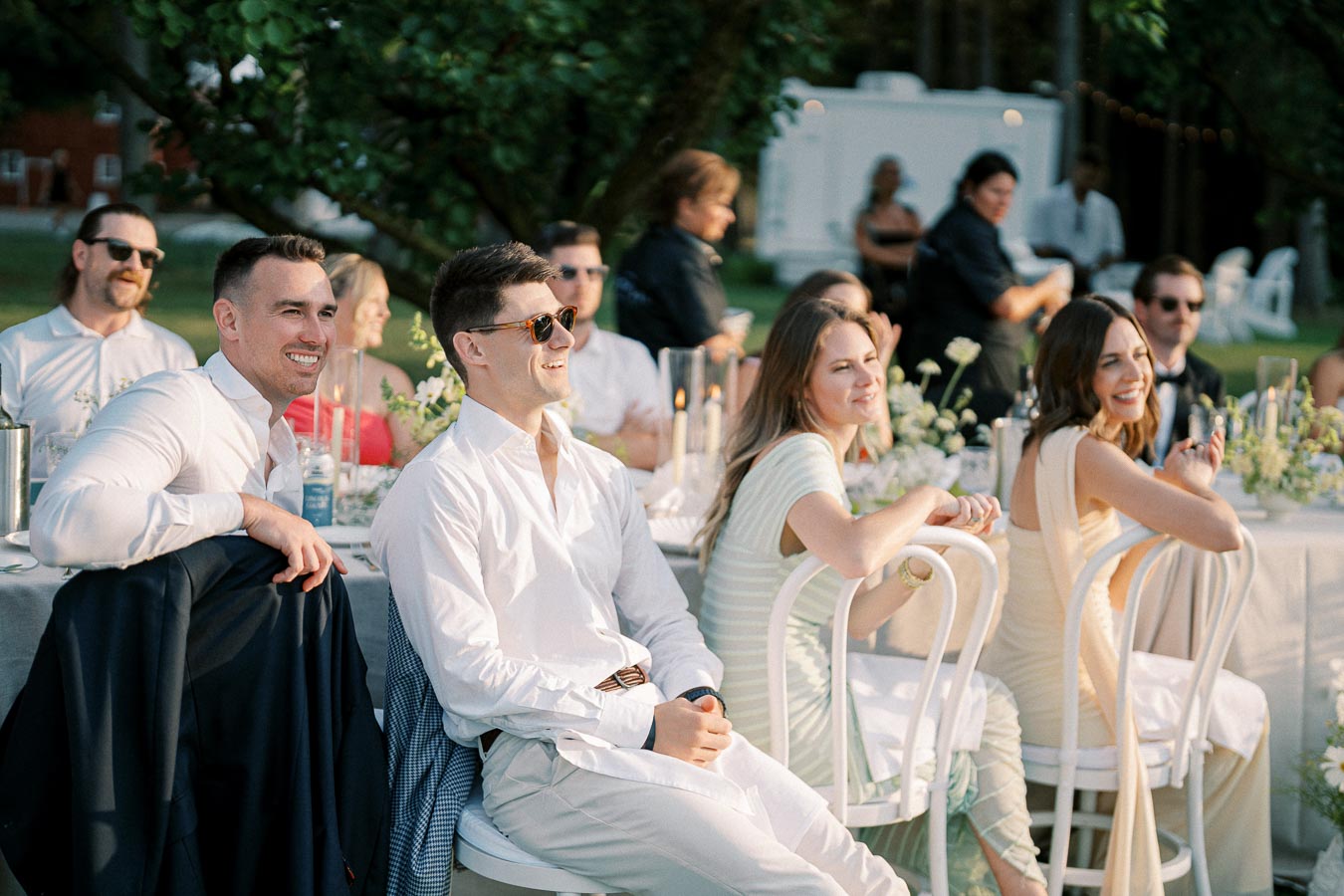 Guests enjoying an outdoor wedding reception, seated at elegantly decorated tables with white chairs and floral centerpieces, under the warm glow of the setting sun.