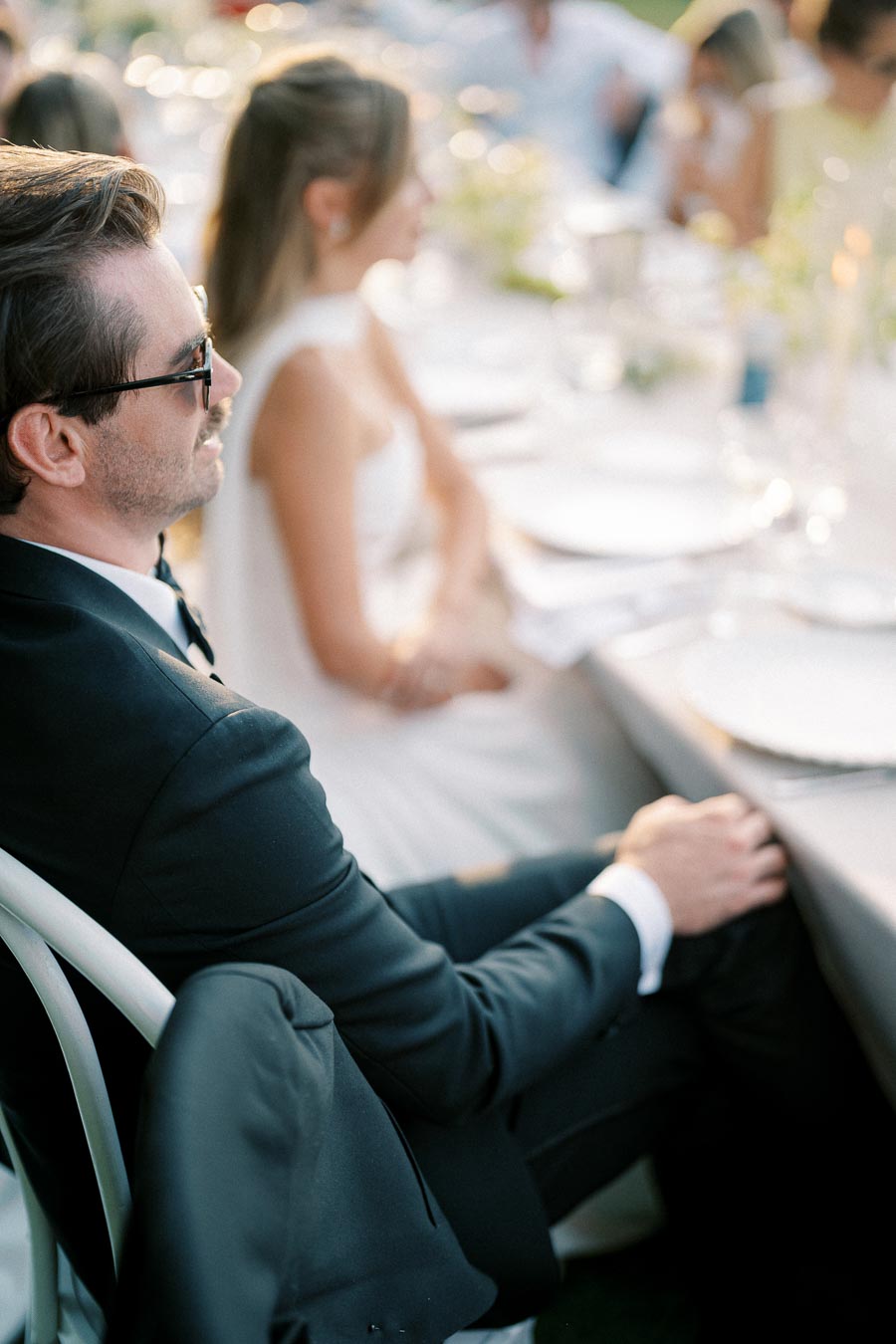 Elegant outdoor wedding reception with guests seated at a beautifully set table, featuring a man in a suit and sunglasses and a woman in a white dress.