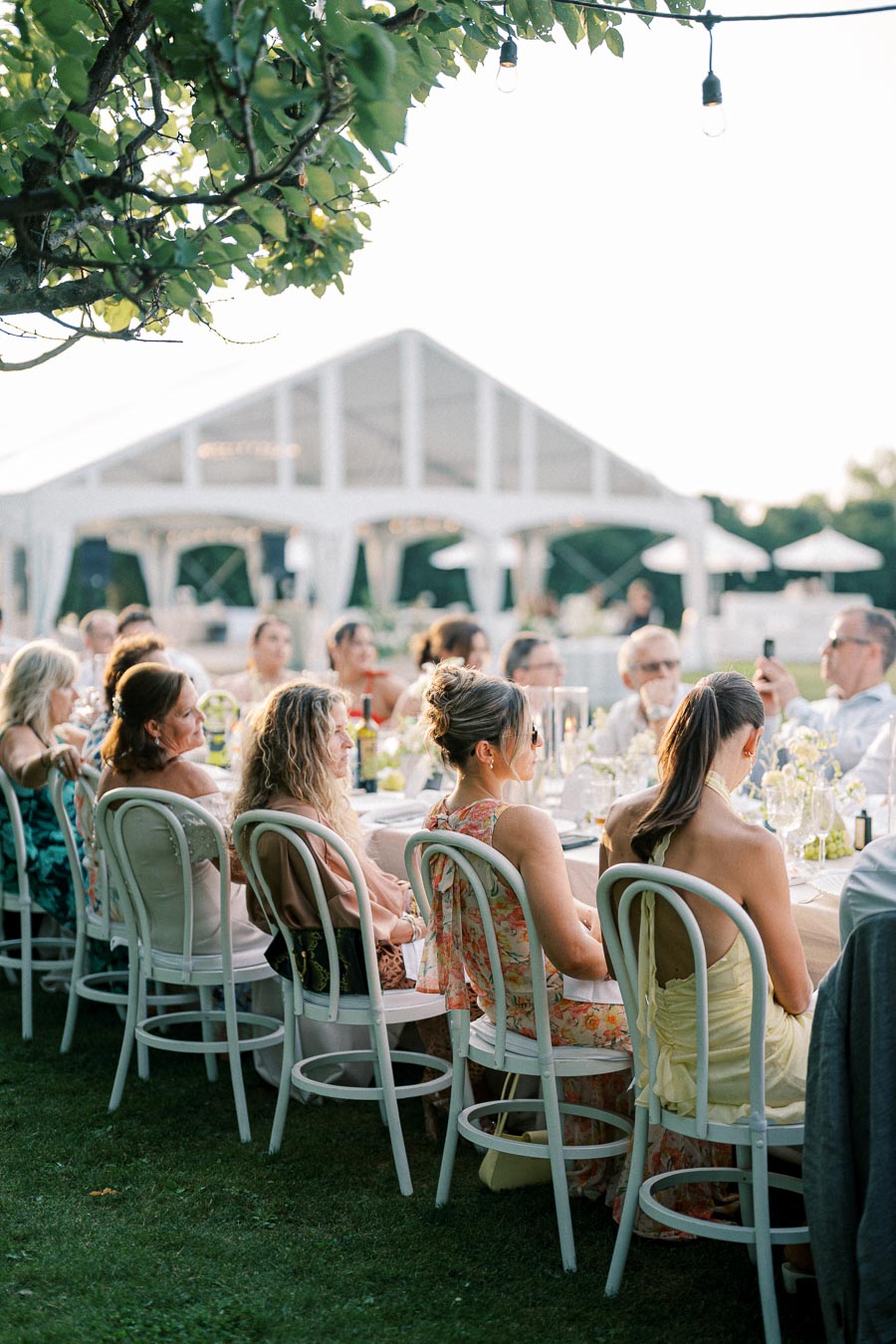 Outdoor wedding reception with elegantly dressed guests seated at a long table under a tent, surrounded by lush greenery and decorative string lights.