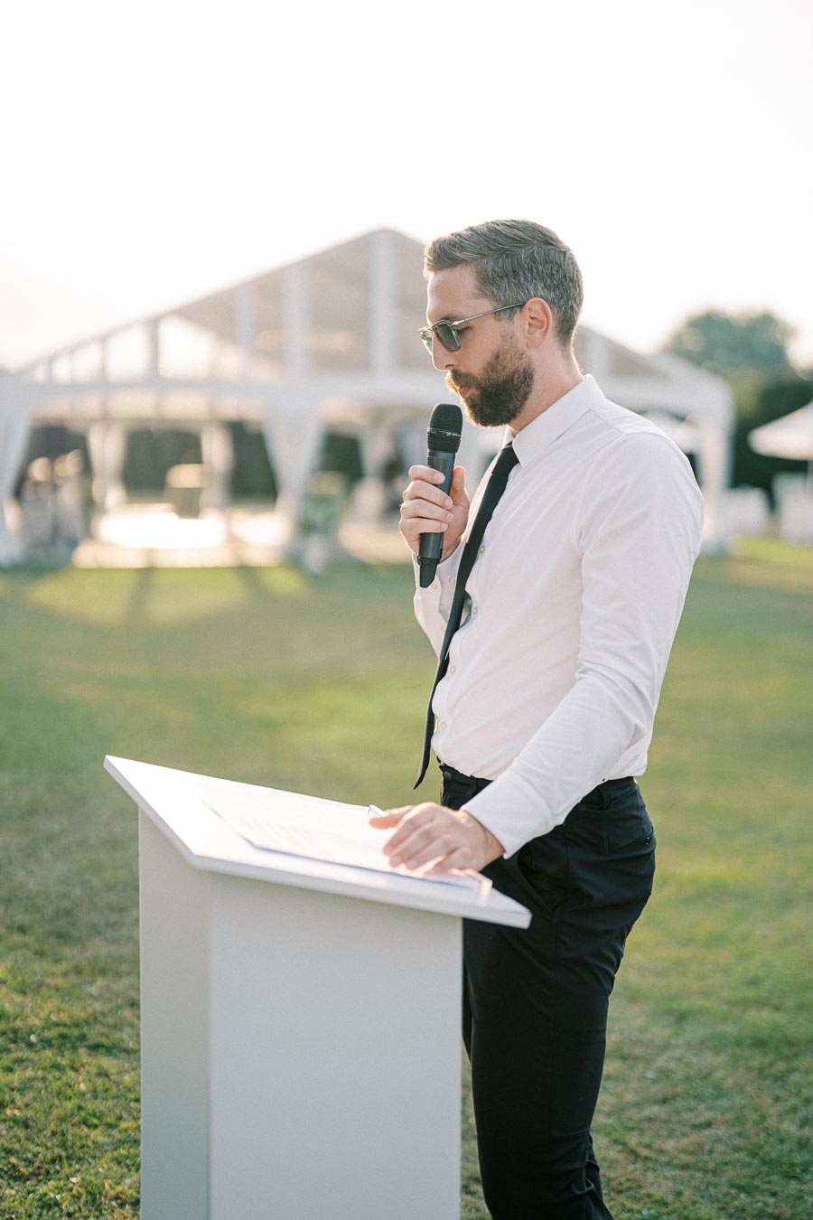 Man in white shirt and black tie delivering a speech outdoors, holding a microphone, with a white event tent visible in the background.