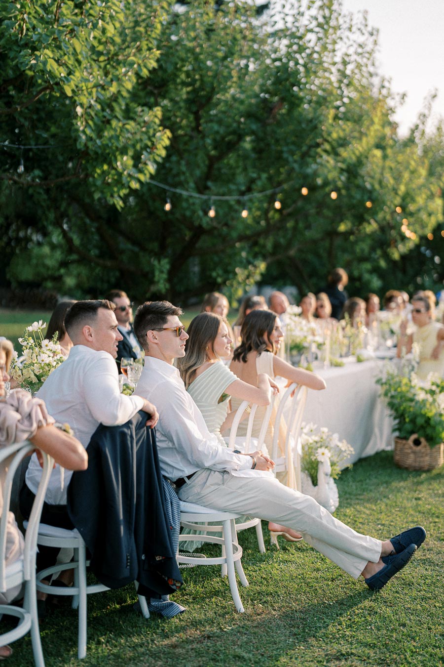 Outdoor wedding reception with guests seated at elegantly decorated tables under string lights, surrounded by lush greenery, enjoying a sunny day celebration.