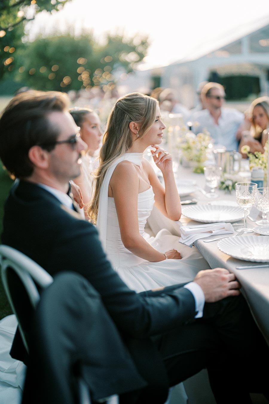 Elegant outdoor wedding reception with guests seated at a beautifully decorated table, featuring white tableware, floral arrangements, and ambient lighting in a garden setting.