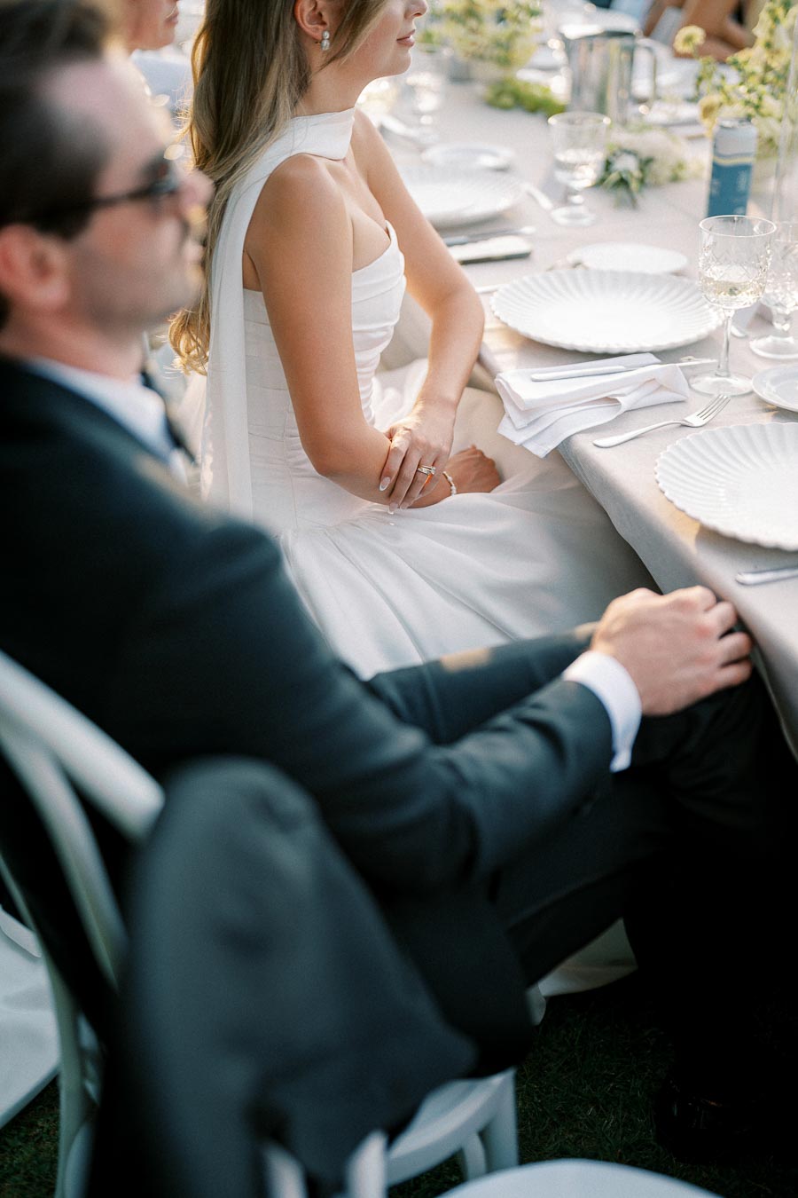 A bride in an elegant white dress sits at a beautifully set wedding reception table, surrounded by guests in formal attire, capturing a serene moment at an outdoor celebration.
