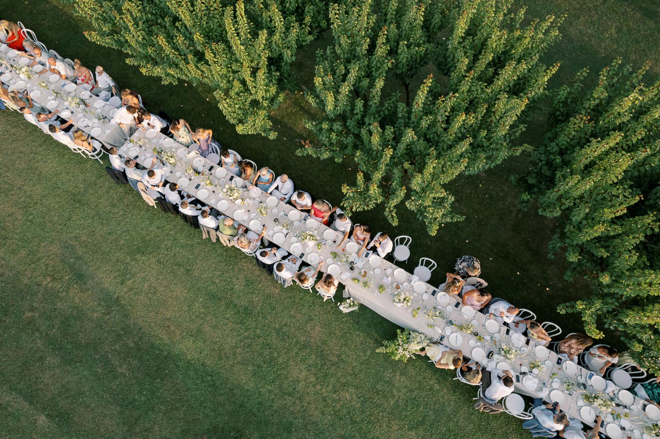 Aerial view of a long outdoor dining table set up for a gathering, surrounded by lush green trees and grass. The table is elegantly arranged for an event, with people seated and enjoying the occasion.