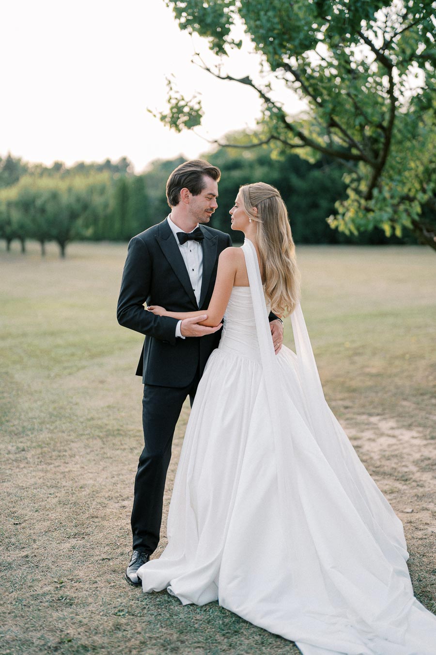 A bride in a flowing white wedding dress embraces a groom in a black tuxedo outdoors, standing on a grassy field with trees in the background.