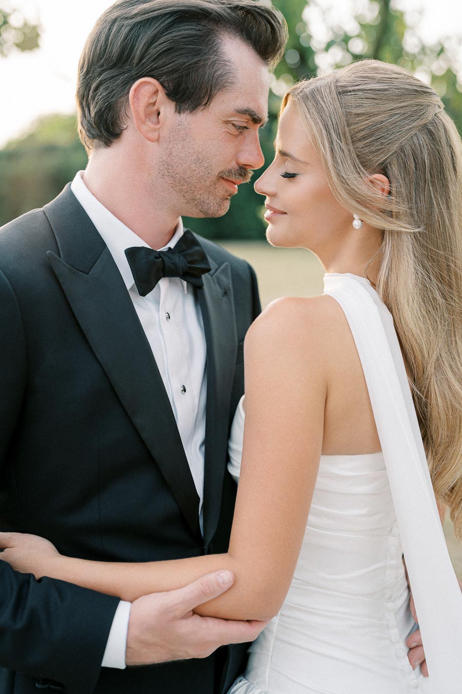 A bride and groom gaze into each other's eyes, embracing in a romantic outdoor wedding setting, with the groom in a classic black tuxedo and the bride in an elegant white gown.