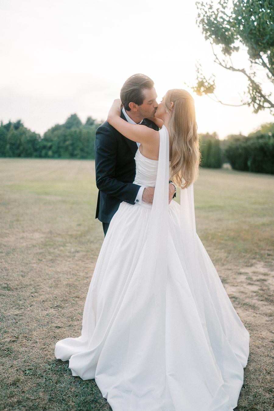 Romantic outdoor wedding couple kissing at sunset, bride in flowing white dress and groom in black suit, surrounded by lush greenery