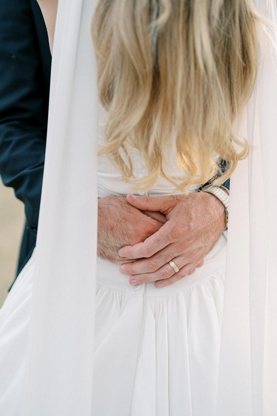 A groom lovingly embraces his bride, showcasing a ring and watch, with her long blonde hair cascading over a white wedding dress.