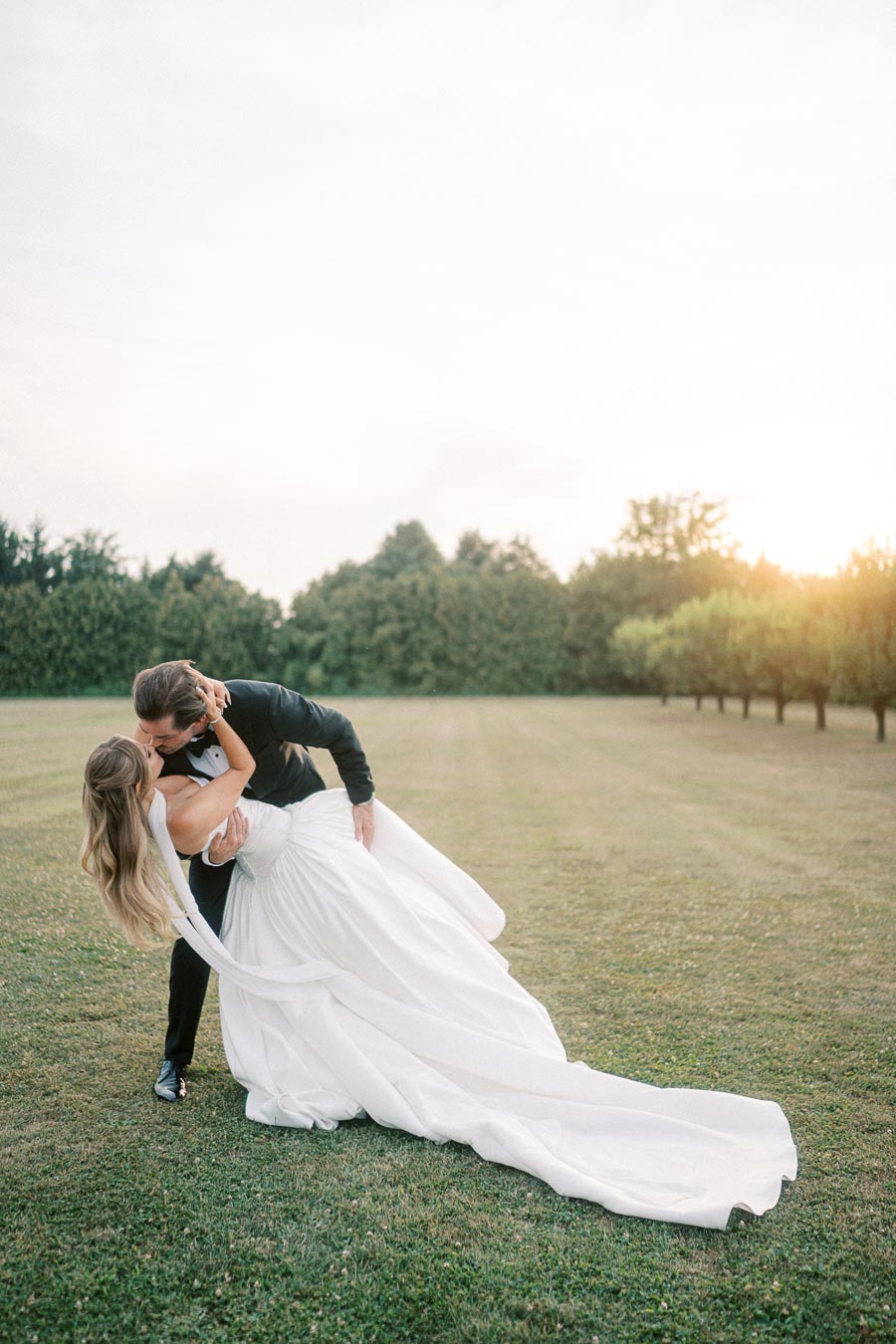 Couple in formal attire sharing romantic kiss outdoors against scenic sunset backdrop in open field.