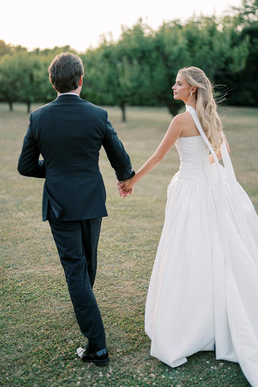 Bride and groom holding hands walking through a grassy field, with the bride in an elegant white dress and the groom in a black suit, surrounded by lush green trees.