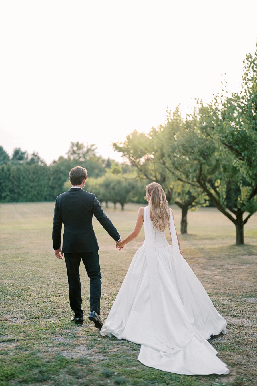 A bride and groom holding hands and walking through a scenic orchard at sunset, with the bride in a flowing white gown and the groom in a classic black suit.