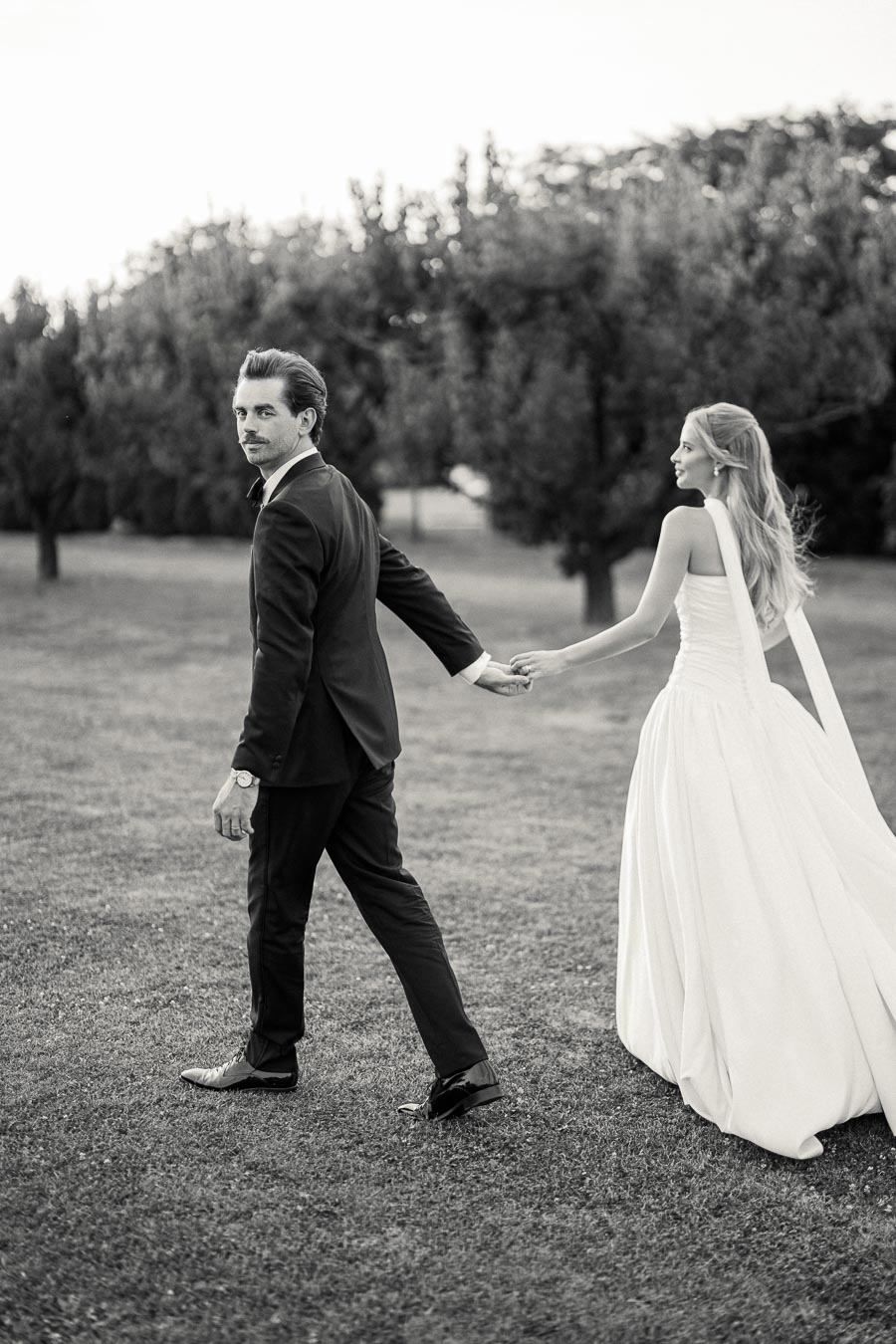 A black and white photograph of a bride and groom walking hand in hand through a grassy field, with the groom in a dark suit looking back and the bride in a flowing white gown smiling, capturing a romantic wedding moment in an outdoor setting.