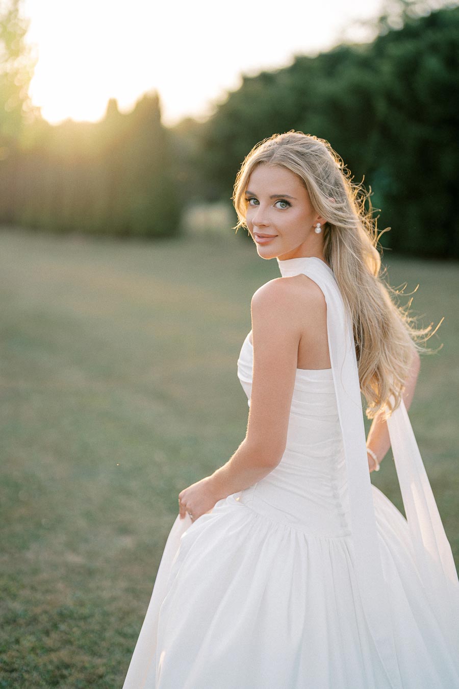 Bride in elegant white gown poses outdoors during sunset, showcasing long flowing blonde hair and serene expression.