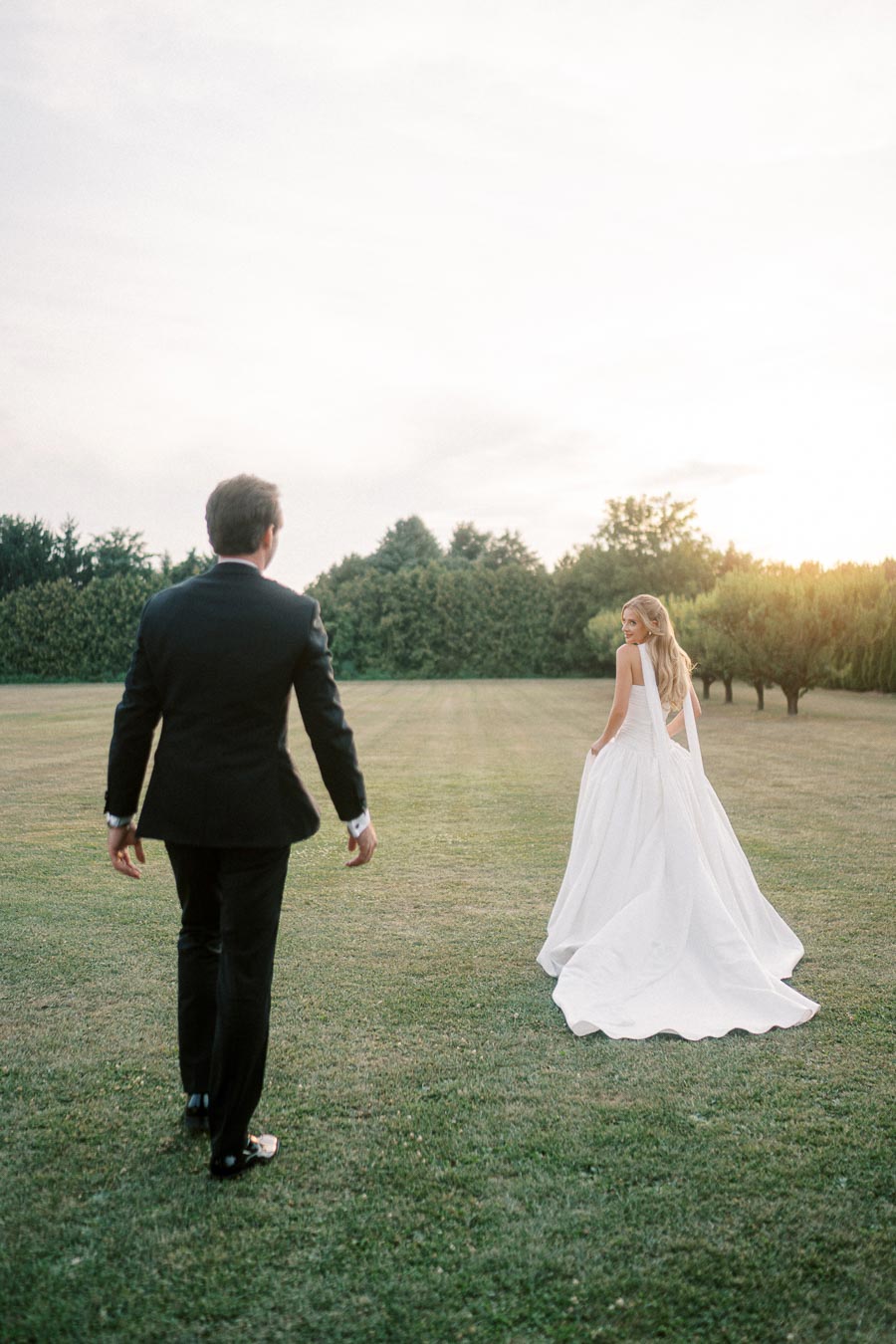 A bride in a flowing white gown walks away, looking back towards a groom in a black suit on a green lawn, surrounded by trees under a soft, cloudy sky.