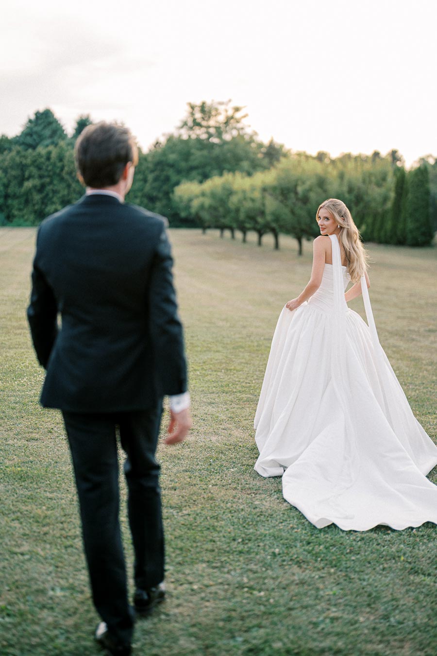 A bride in a flowing white wedding dress walking on a grassy field, gazing back with a smile, while a groom in a black suit approaches, surrounded by lush greenery and trees.