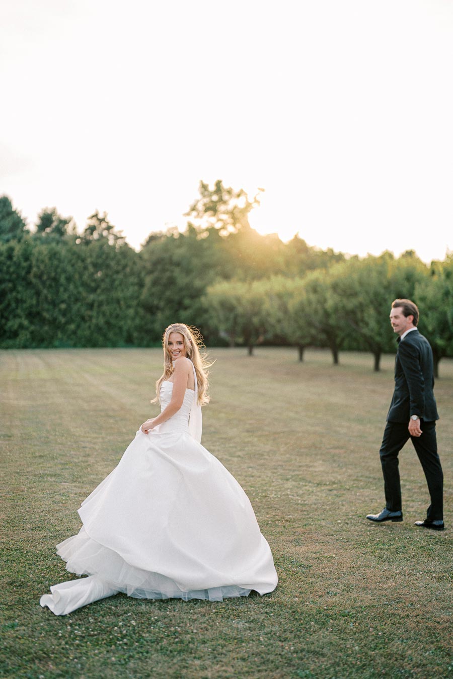 A bride in a flowing white wedding dress smiles joyfully as she walks across a sunlit field with a groom in a black suit following, surrounded by lush greenery.
