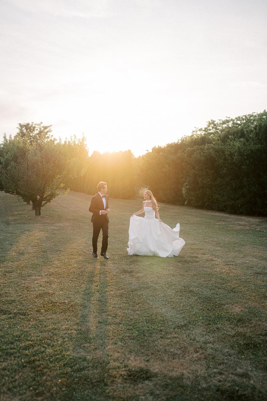 Sunset wedding photo of a bride in a flowing white gown and groom in a black tuxedo, walking in a sunlit garden with green trees in the background.