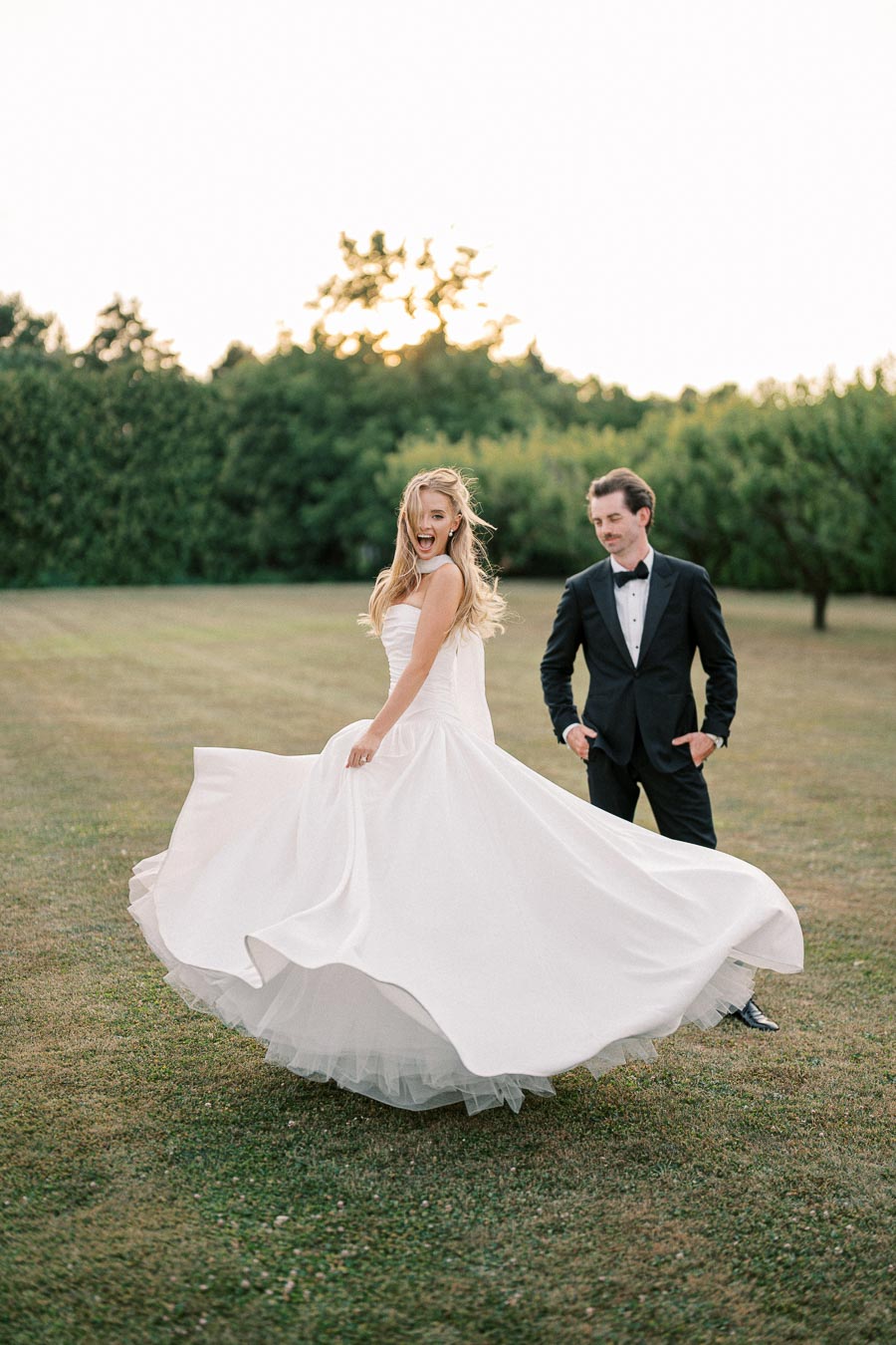 A bride in a flowing white dress twirls joyfully on a grassy field, with a groom in a tuxedo smiling in the background, set against a backdrop of lush green trees at sunset.