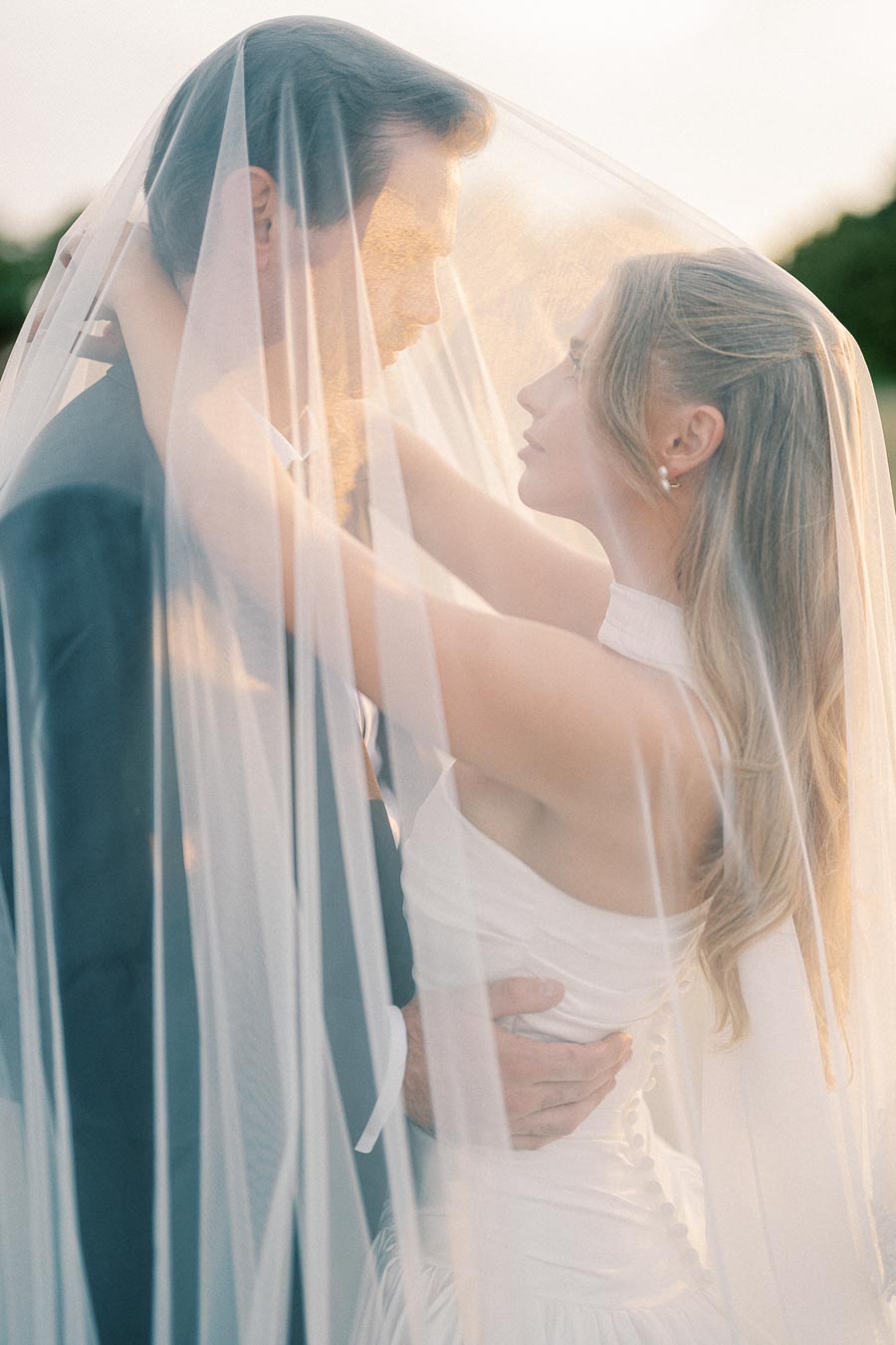 Romantic bride and groom embracing under a flowing veil during sunset, capturing a serene and intimate wedding moment.