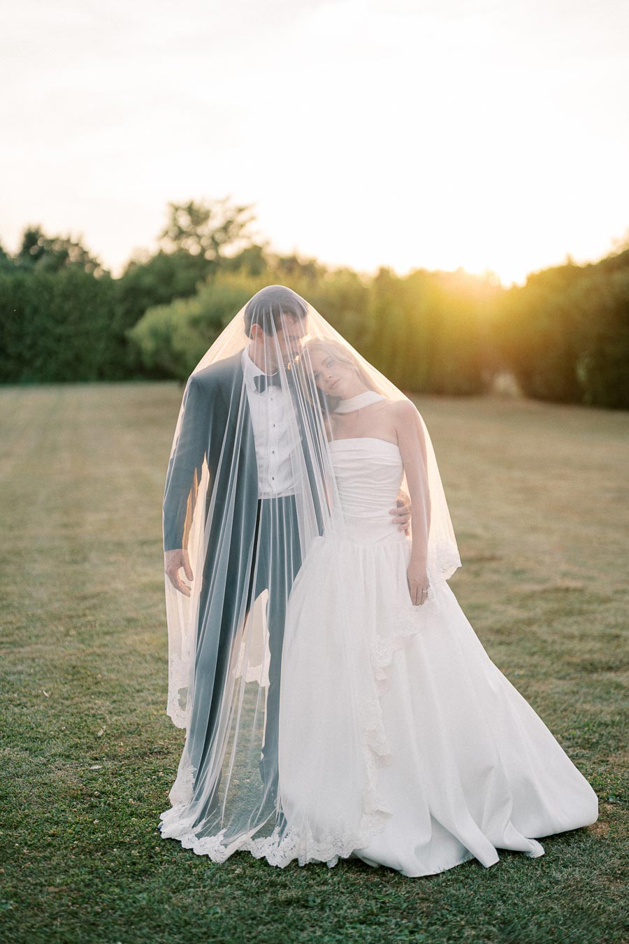Romantic outdoor wedding scene with a bride and groom embracing under a sheer veil, set against a serene grassy field at sunset.