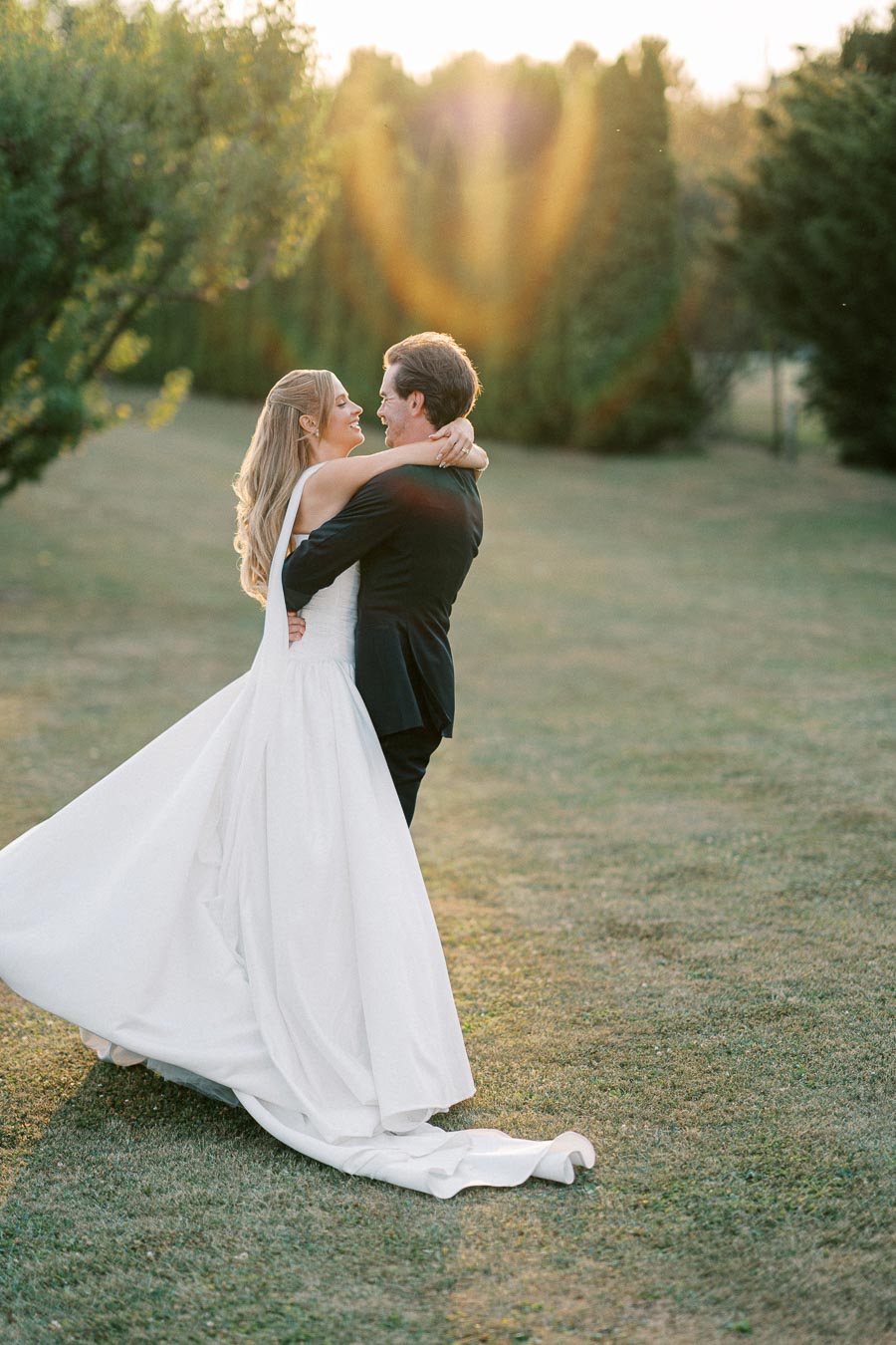 A couple embracing each other on their wedding day, with the bride in a flowing white gown and the groom in a black suit, standing on a lush green lawn, as the sun sets behind them, casting a warm glow.