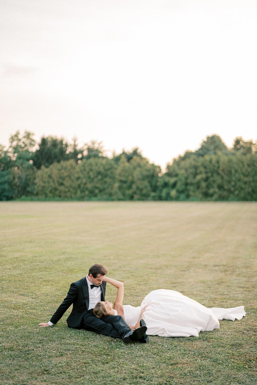 A bride in a white gown and groom in a black tuxedo relax on a grassy field, surrounded by greenery, under an open sky.