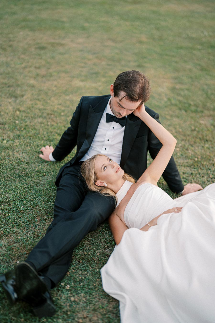 A romantic moment with a bride in a white wedding dress lying on a groom's lap in a tuxedo on a grassy field, showcasing their love on a picturesque wedding day.