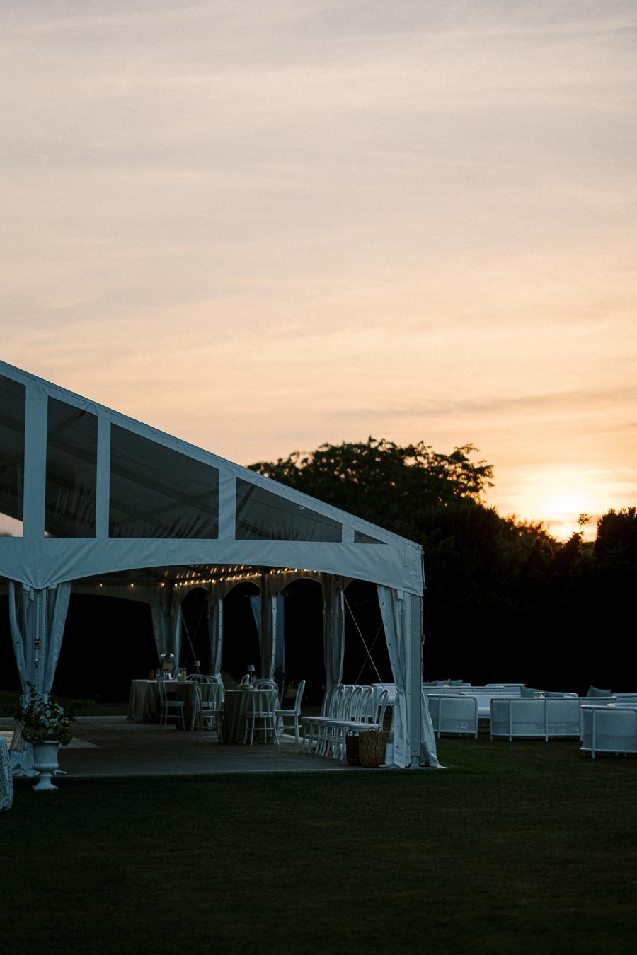 Outdoor wedding tent setup at sunset with elegant table settings and chairs, creating a serene and romantic atmosphere.