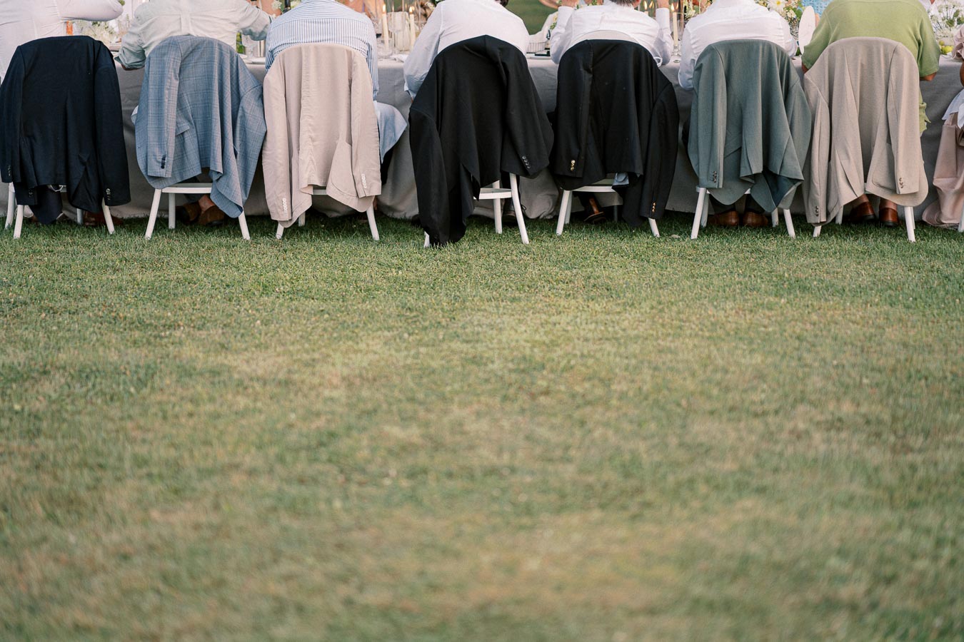 Wedding guests sitting at a long outdoor table, with jackets draped over chairs and a grassy lawn in the foreground.
