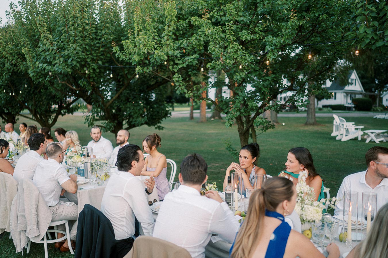 Outdoor wedding reception with elegantly dressed guests seated at a long dining table, surrounded by lush greenery and ambient lighting.