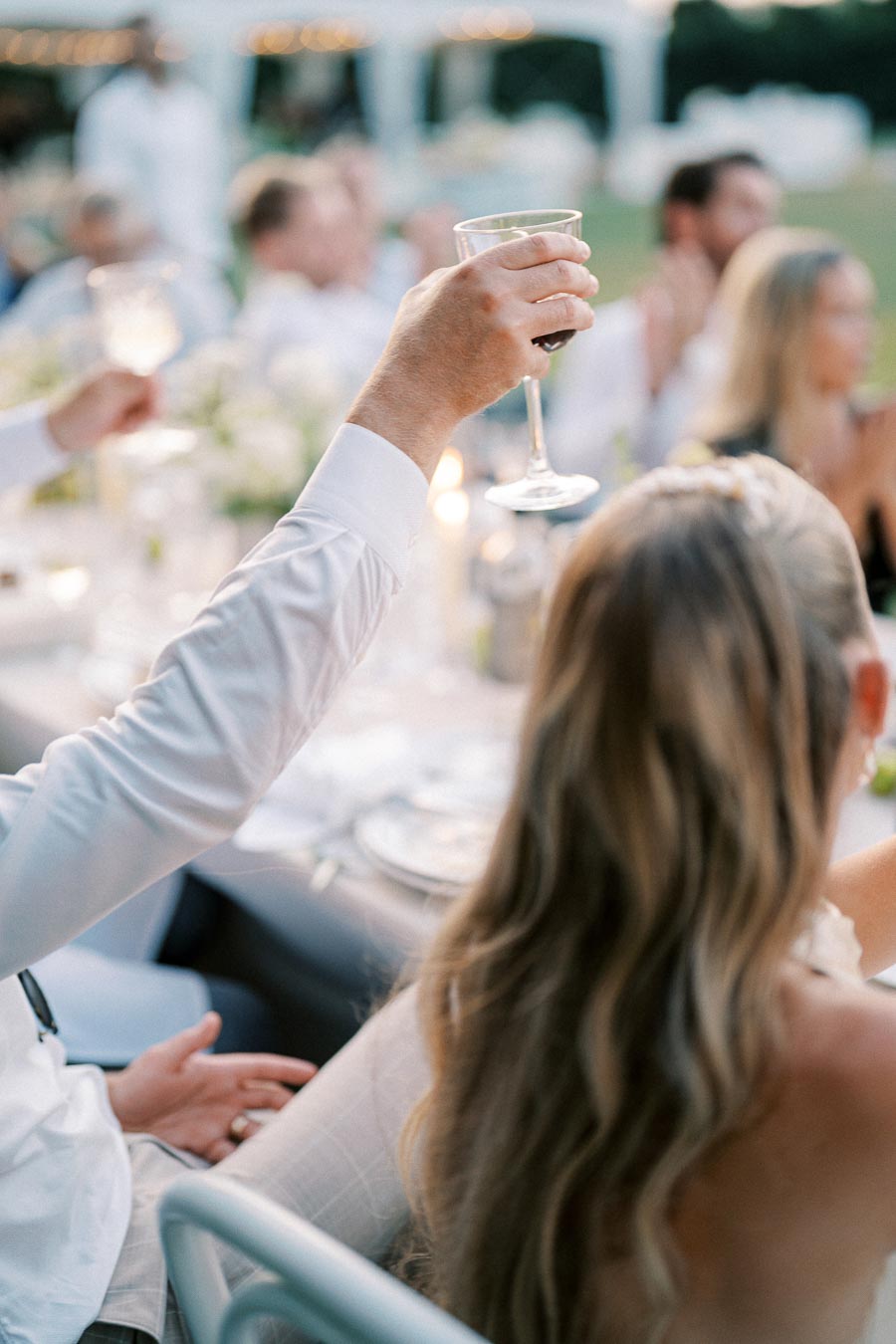 A group of elegantly dressed guests raising wine glasses for a toast at an outdoor wedding reception, celebrating with white flowers and candlelit table settings.