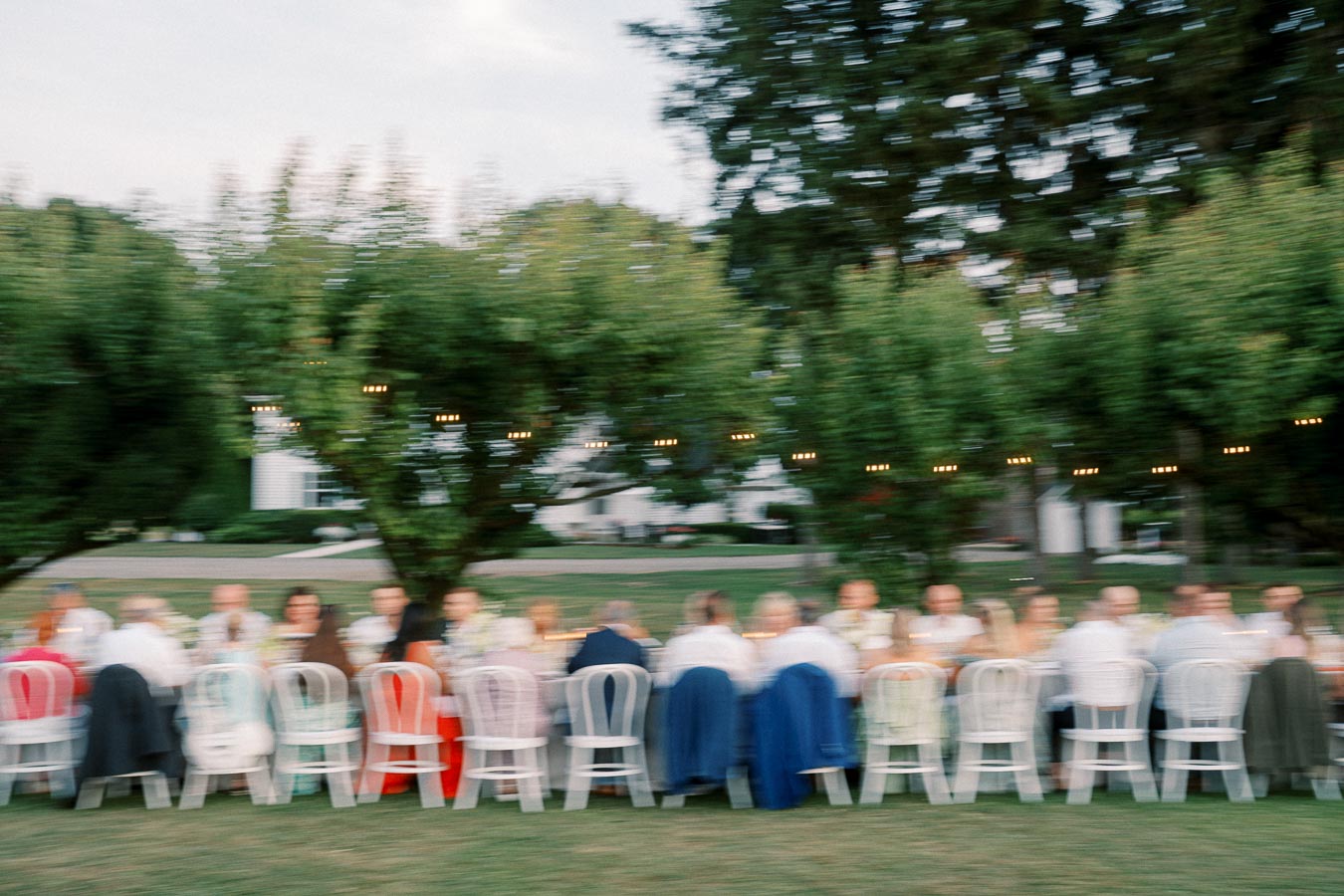 Blurred image of a large group of people sitting at a long outdoor dining table during an event, surrounded by greenery and string lights, evoking a warm and festive atmosphere.