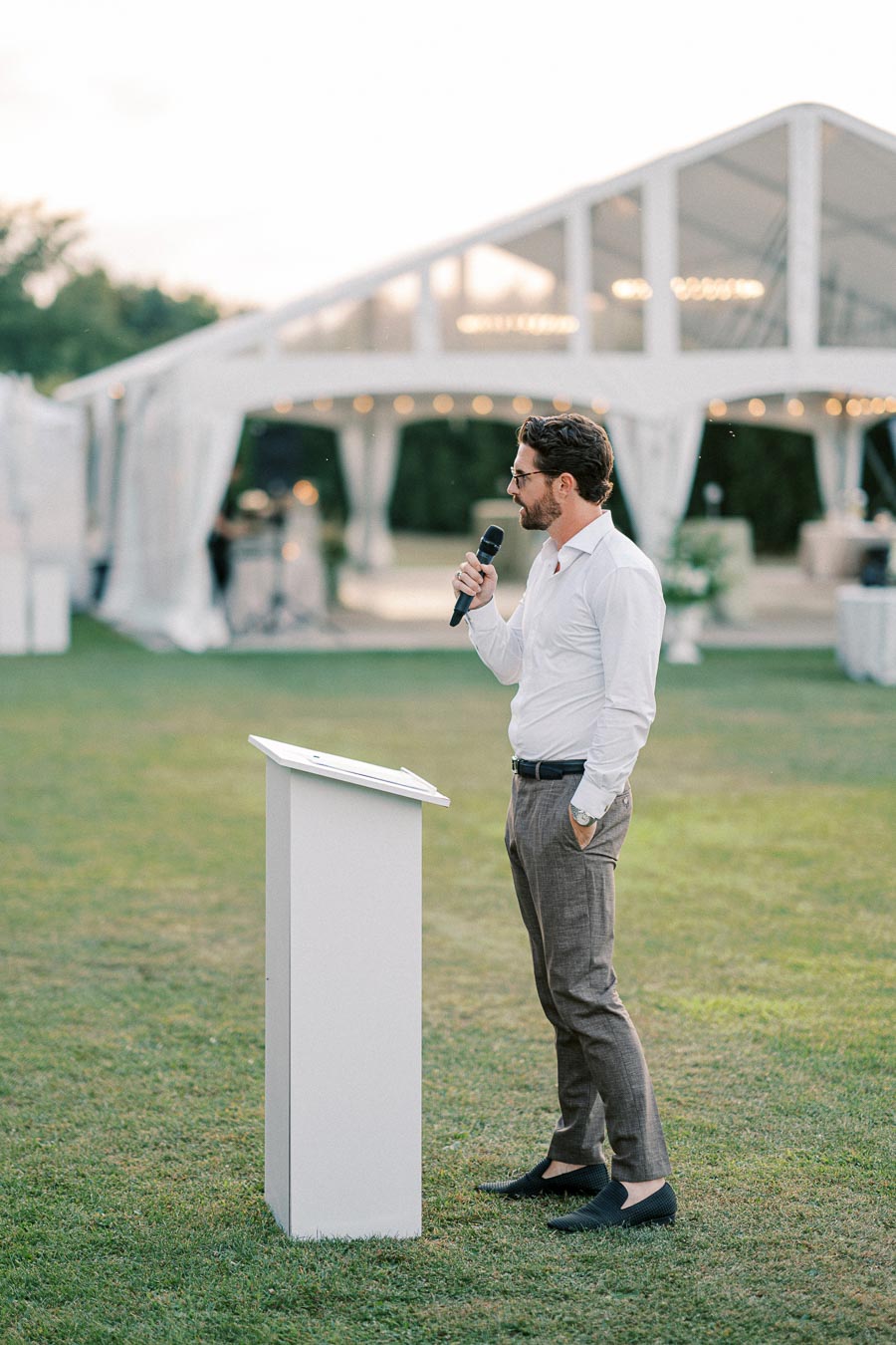 A man in formal attire giving a speech outdoors at an elegant event, standing at a podium with a microphone, with a white tent and greenery in the background.