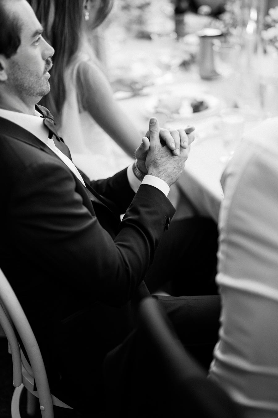Black and white photo of a man in a suit sitting at a formal dining table, hands clasped, focused and contemplative, during an elegant gathering or event.