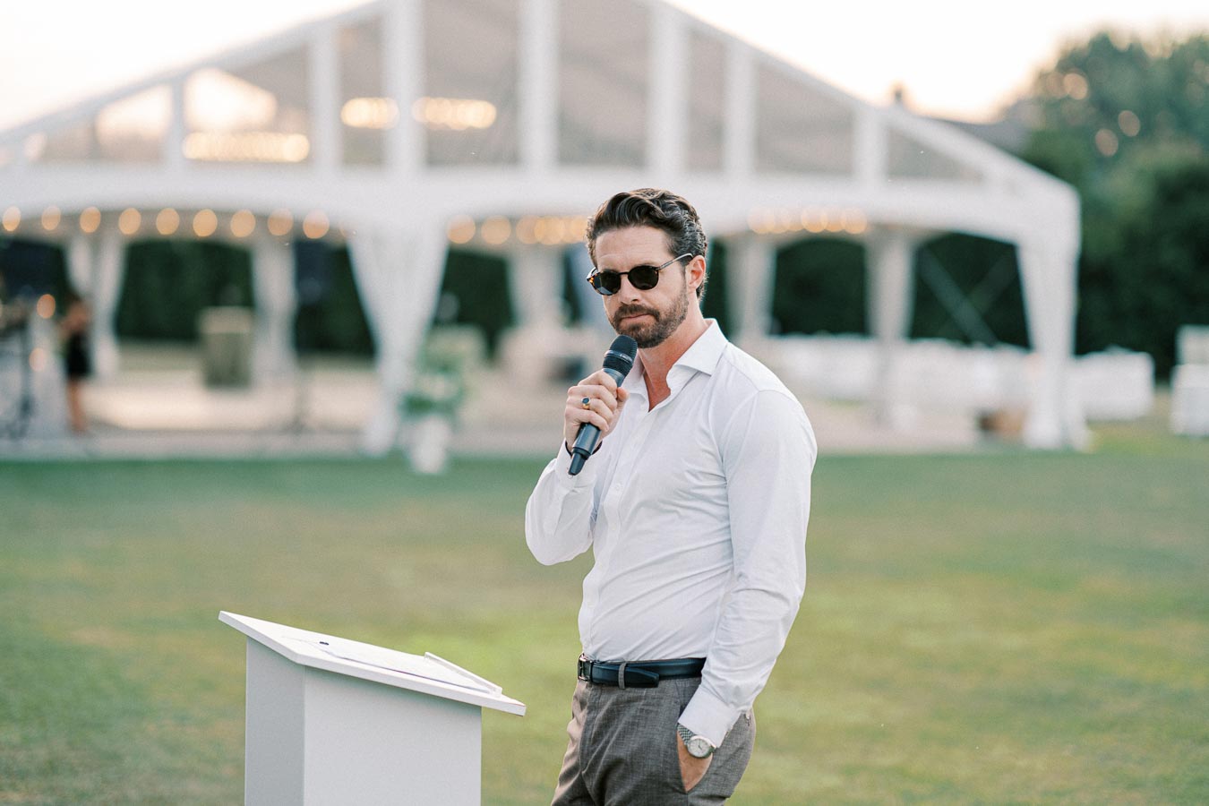 Man in sunglasses speaking into a microphone at an outdoor event, standing next to a podium with a white tent in the background.
