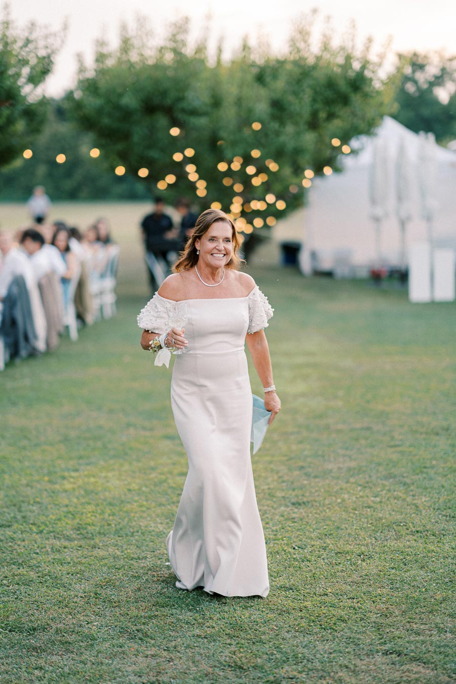 A woman in a white, off-the-shoulder dress walks on a grassy lawn during an outdoor event, holding a wine glass, with string lights and blurred people in the background.