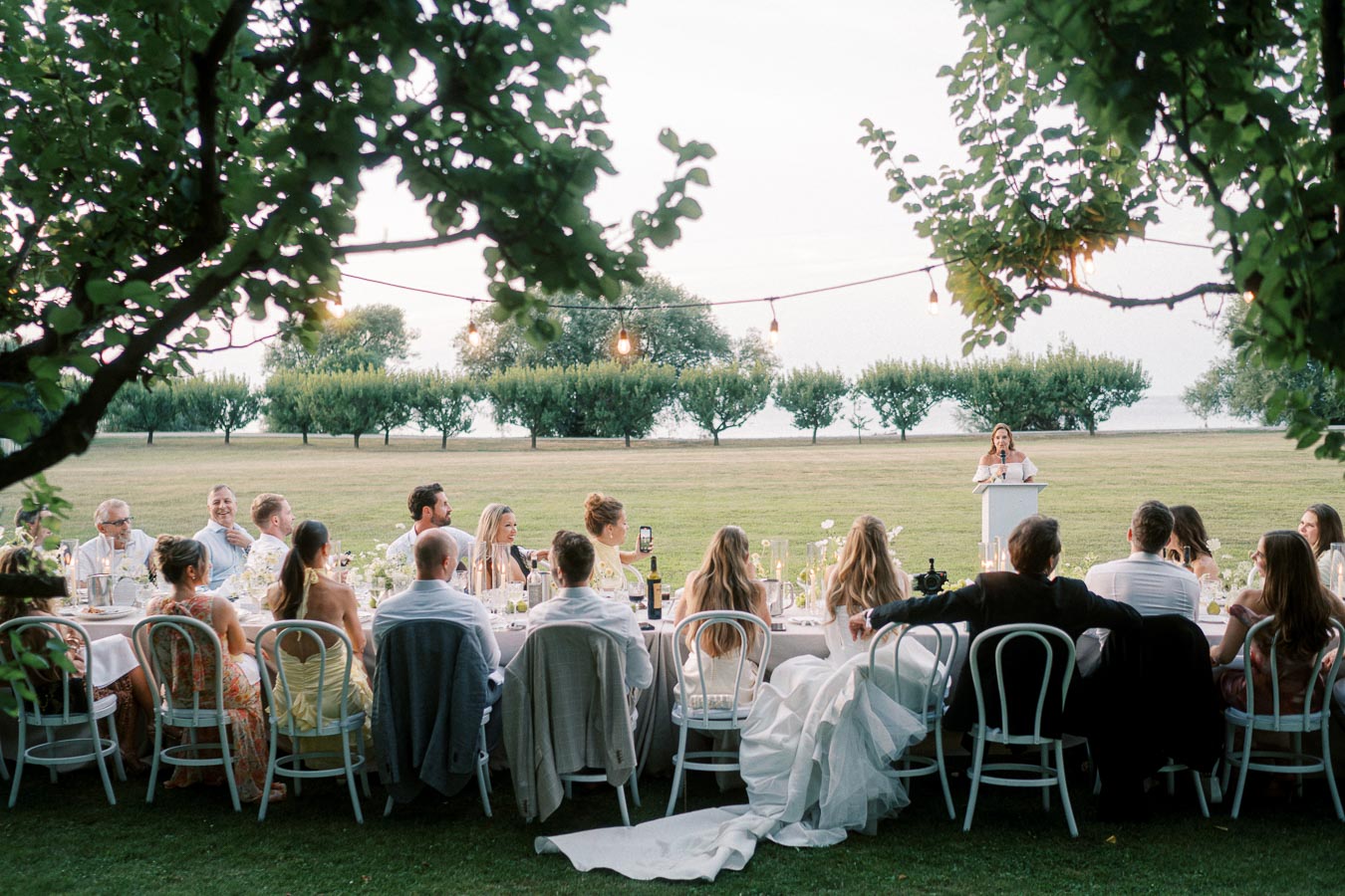 Outdoor wedding reception with guests seated at a long table beneath string lights, listening to a speech on a sunny day.