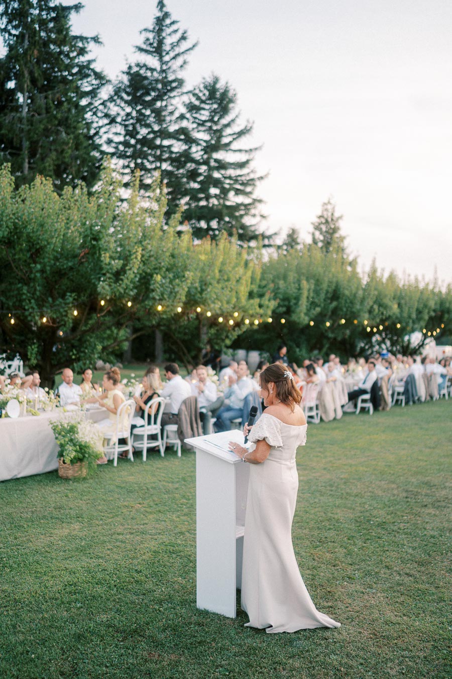 A woman in an elegant white dress giving a speech at a long outdoor reception table, adorned with white flowers and surrounded by guests, set against a backdrop of lush green trees and hanging string lights.