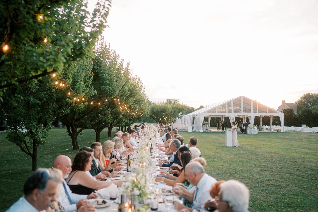 Outdoor wedding reception with a long dining table set on a green lawn. Guests are seated under string lights, enjoying a meal near a white tent during sunset.