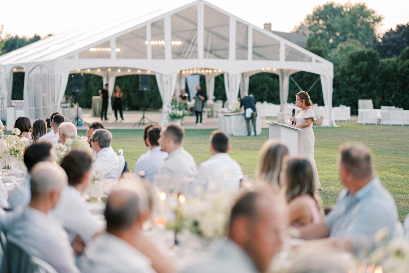 Outdoor wedding reception with guests seated at elegantly decorated tables, a woman giving a speech at a podium, and a large clear tent in the background surrounded by greenery.