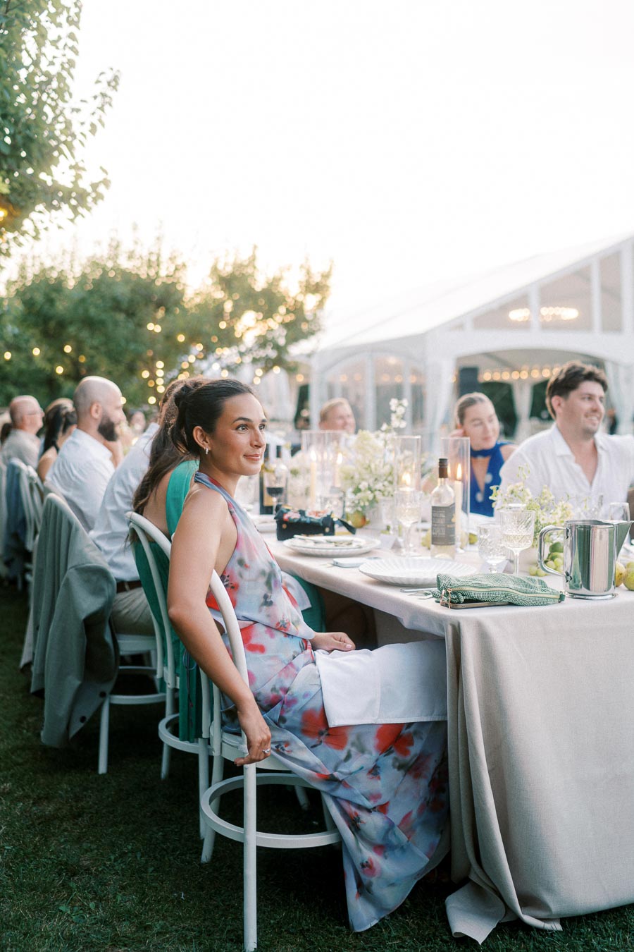 Outdoor wedding reception with elegantly dressed guests seated at a table adorned with floral arrangements and candles, set under a bright sky and surrounded by greenery.