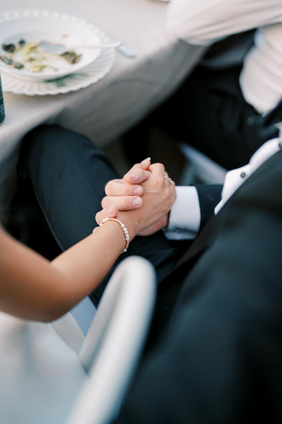 A close-up view of a couple holding hands at a formal event, with a table setting in the background, highlighting the intimacy and connection.