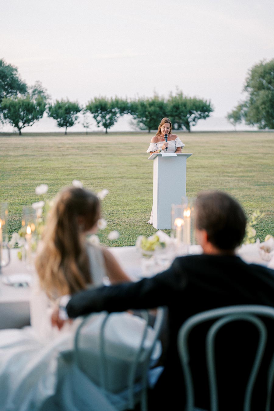 Wedding reception speech outdoors, woman in white dress addressing guests seated at elegantly decorated table, with greenery and ocean in the background.