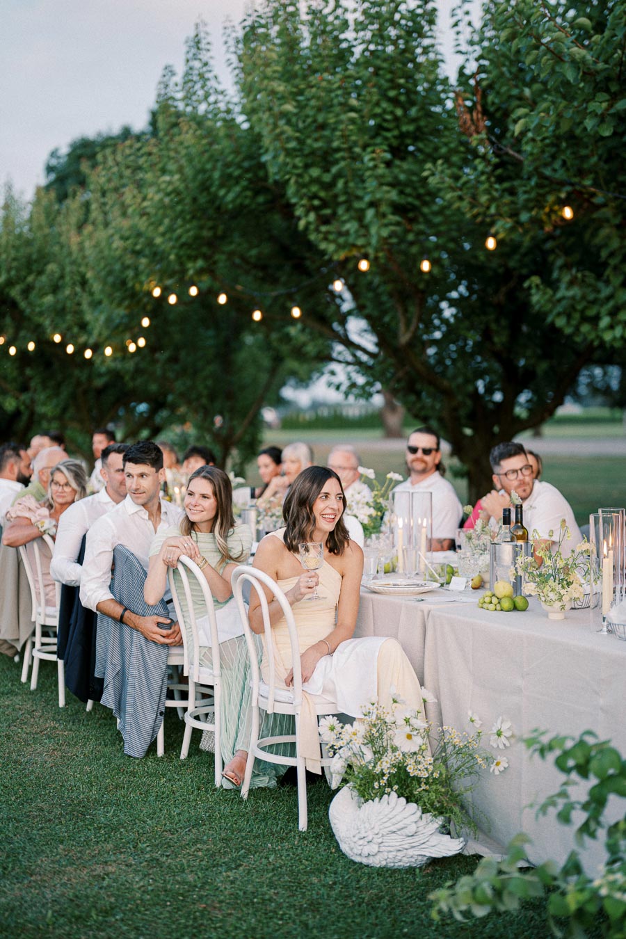 Guests enjoying an elegant outdoor dinner party with string lights and floral decorations, seated at a long, beautifully set table in a lush garden setting.