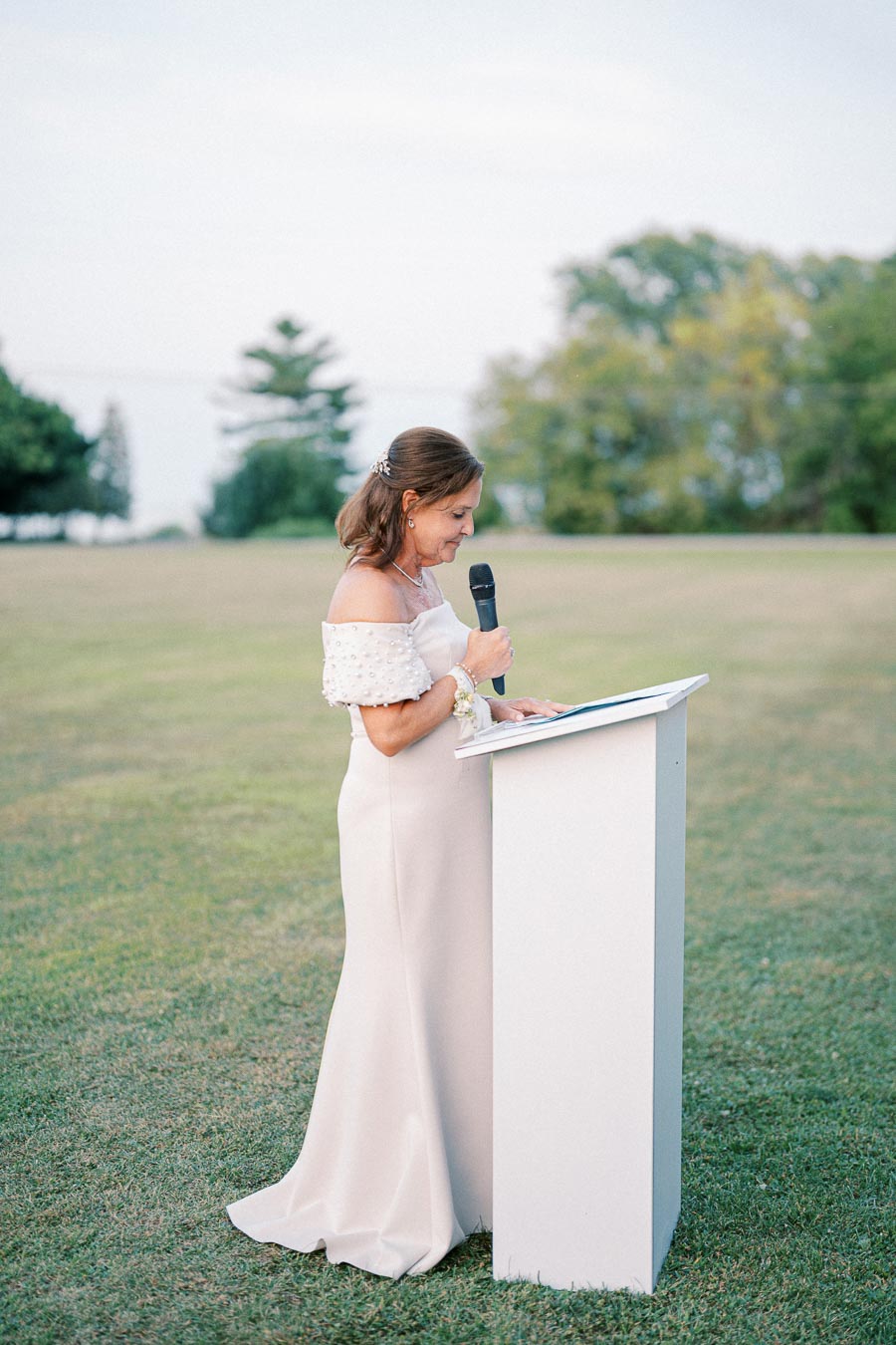 A woman in an elegant off-the-shoulder gown gives a speech outdoors using a microphone while standing at a podium on a grassy lawn, with trees and a slightly overcast sky in the background.