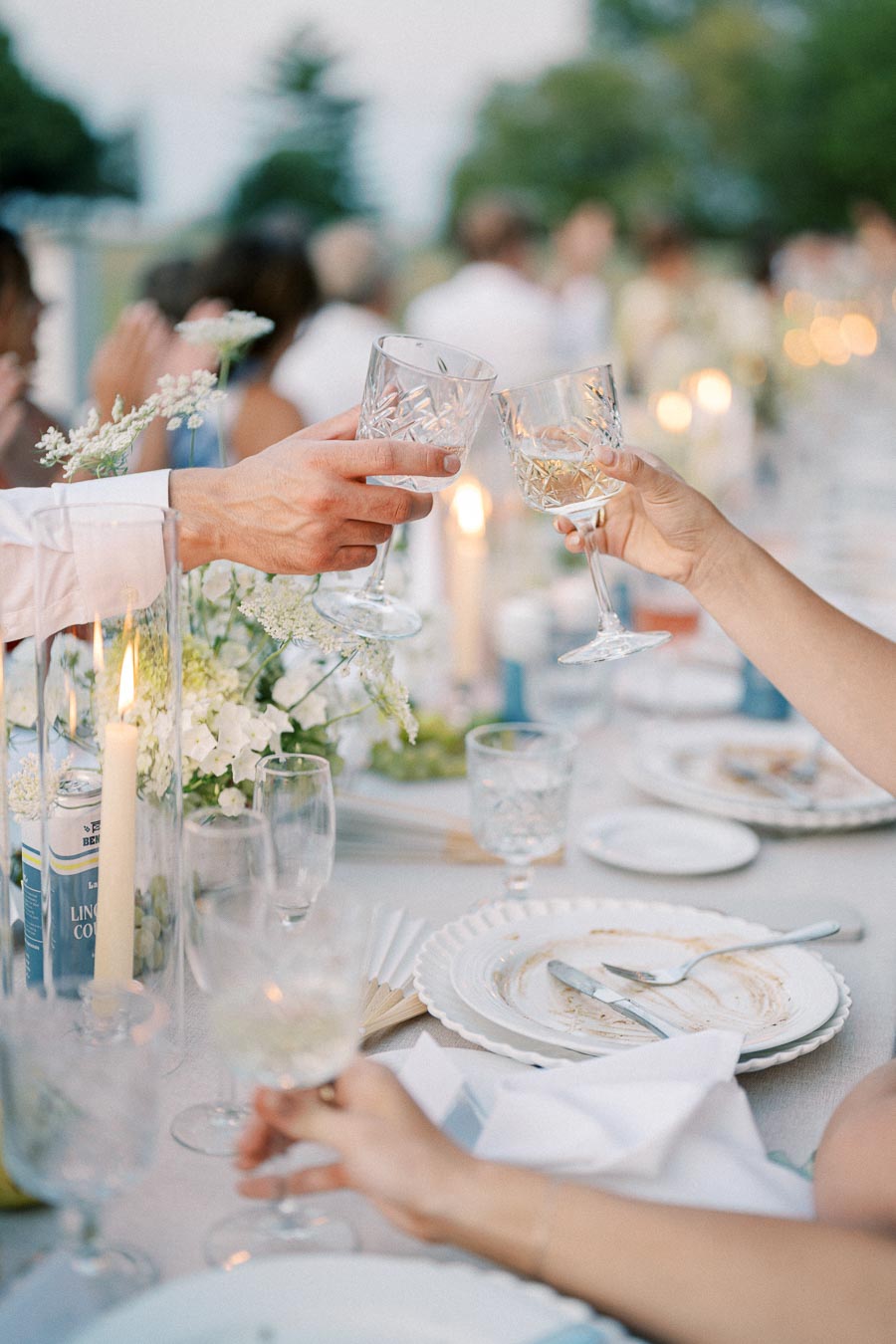 Elegant wedding reception table setting with guests toasting with crystal glasses, surrounded by candlelight and floral arrangements on an outdoor dining table. Celebratory, romantic atmosphere.