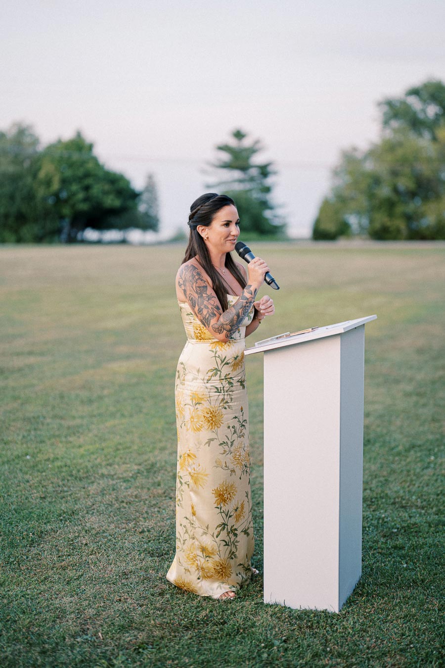 Woman with long dark hair and floral tattoo giving a speech outdoors at a podium, wearing a yellow floral dress against a backdrop of greenery and open sky.