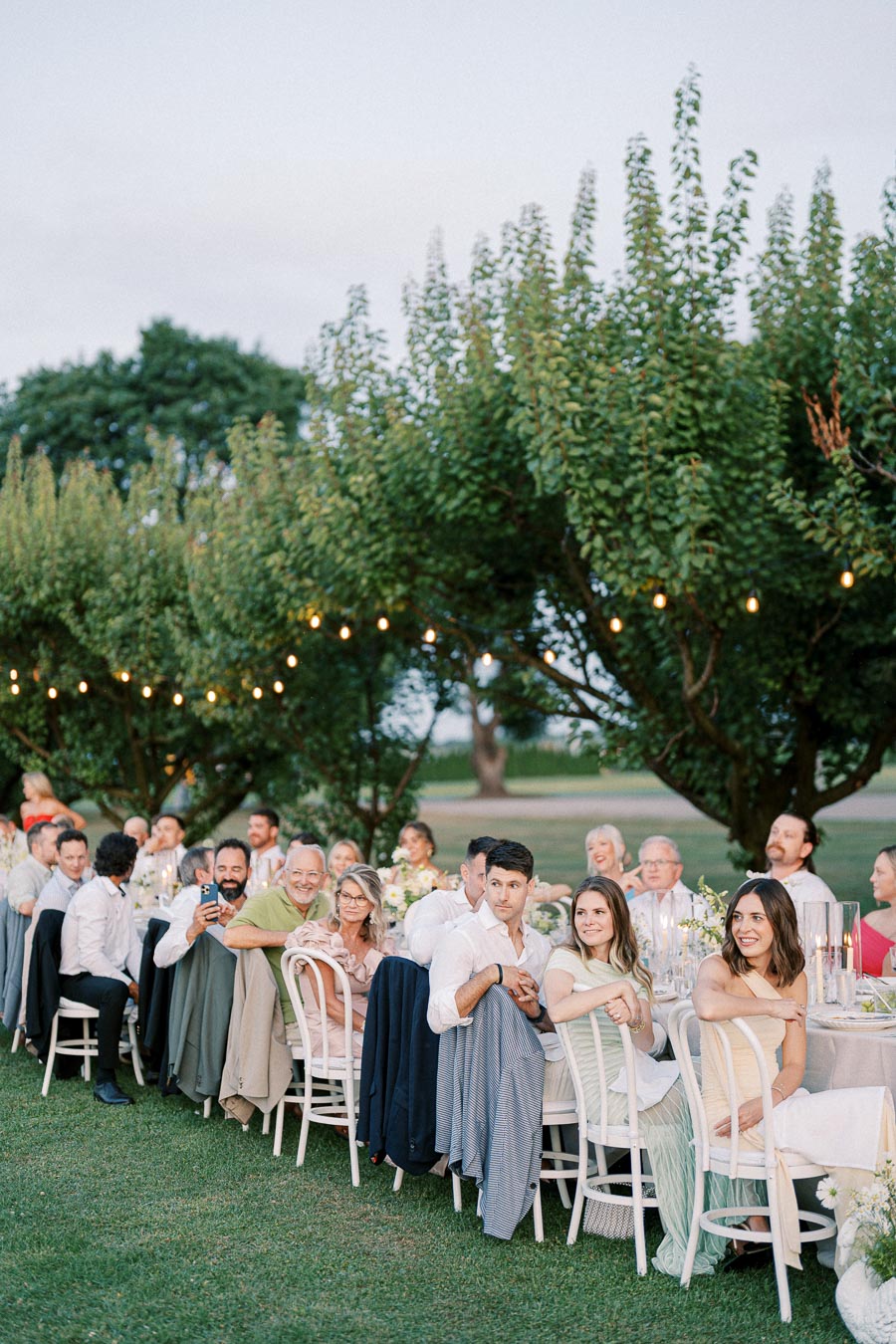 Outdoor wedding reception with guests seated at a long table, surrounded by greenery and string lights, during a beautiful evening.